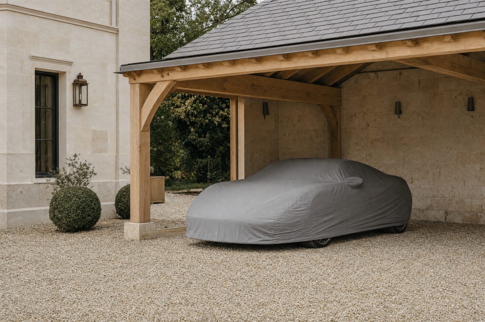 Une voiture de sport au repos sous une housse grise ajustée, garée sous un carport en bois, gravier clair au sol.